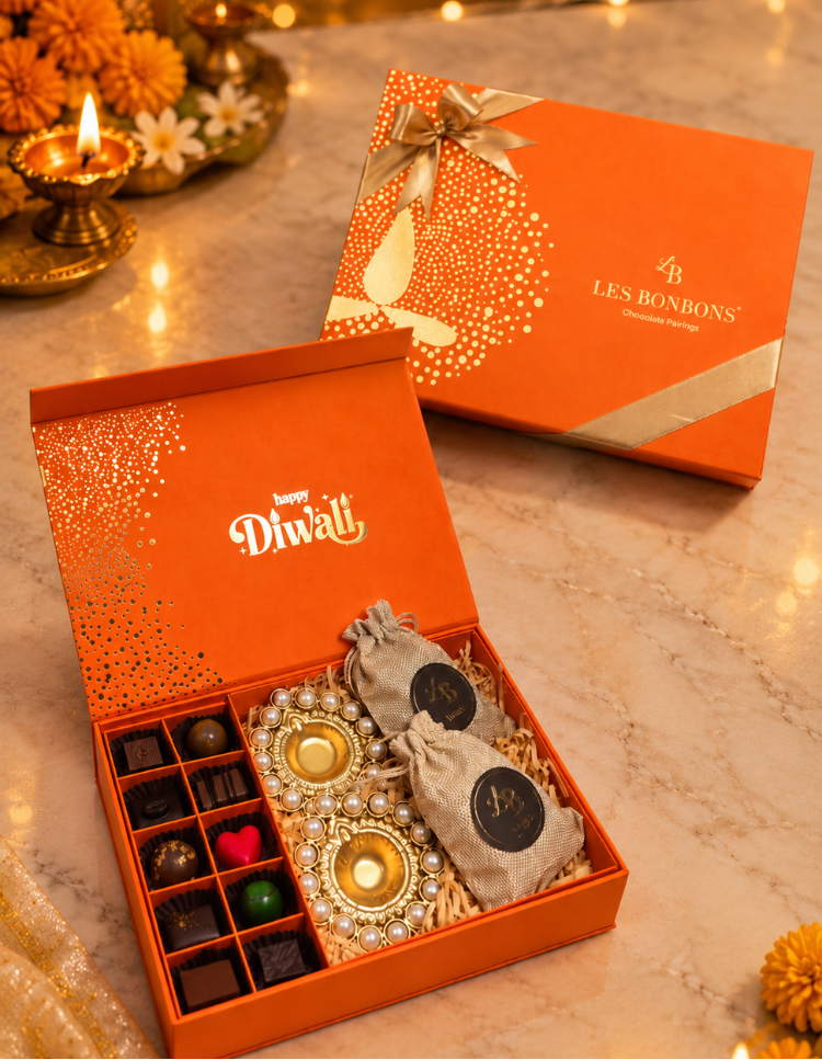An orange Les Bonbons Diwali gift box shown open on a marble surface, with assorted chocolates and decorative sweets inside. The matching closed box sits behind it on the same surface, with warm string lights and candlelight in the background.